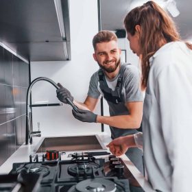 Young professional plumber in grey uniform shows results of work for housewife on the kitchen